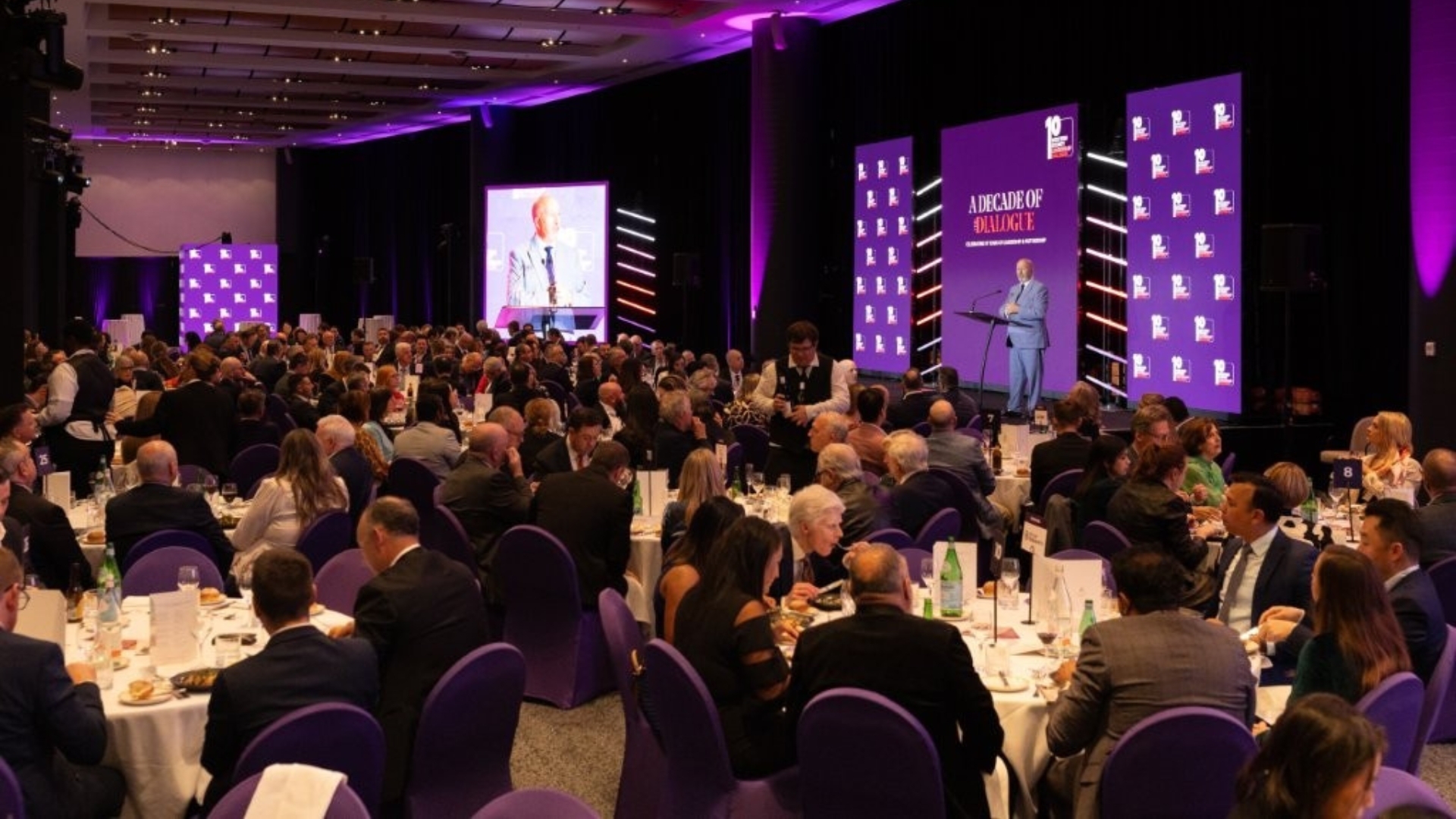 Wide shot of event room with crowd, speaker on stage and LED screen 