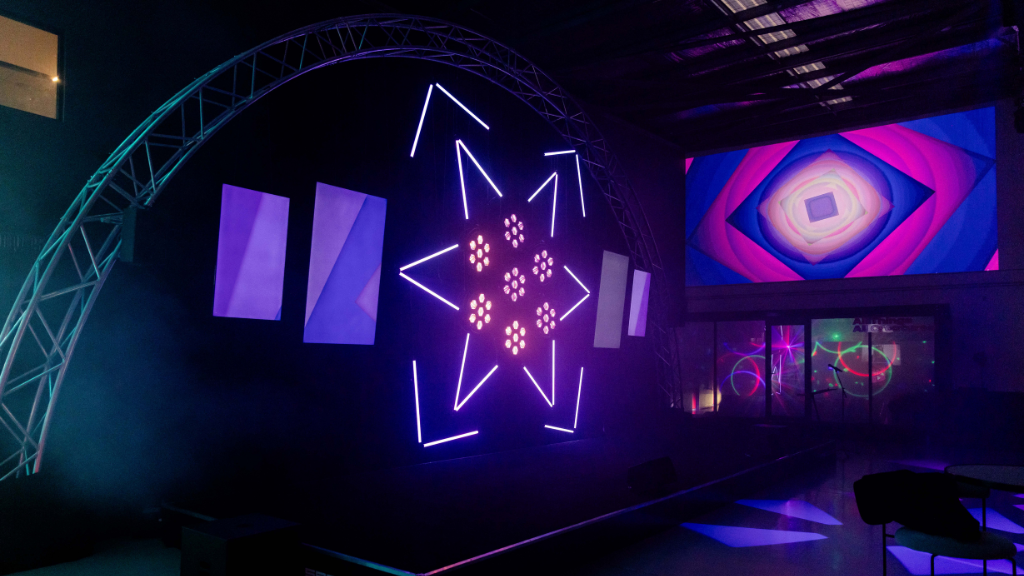 Two televisions hanging on either side of a custom LED light centrepiece in the shape of a star on stage. On the right is a wall projection in a kaleidoscopic pattern.